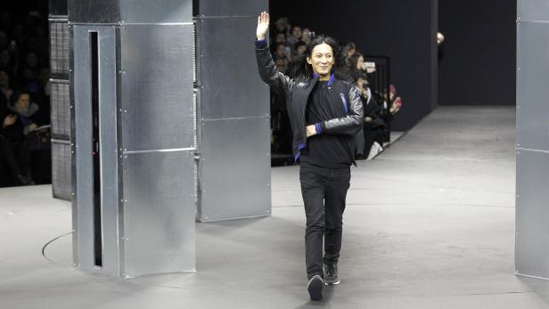 Ein Mann mit langen Haaren winkt auf einem Laufsteg zwischen silbernen Metallstrukturen.