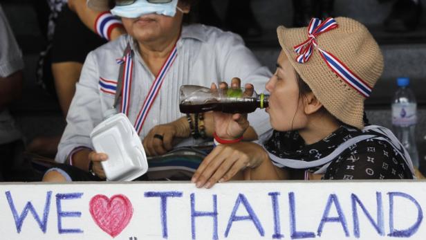 Eine Frau mit Hut trinkt aus einer Flasche vor einem Schild mit der Aufschrift „We love Thailand“.