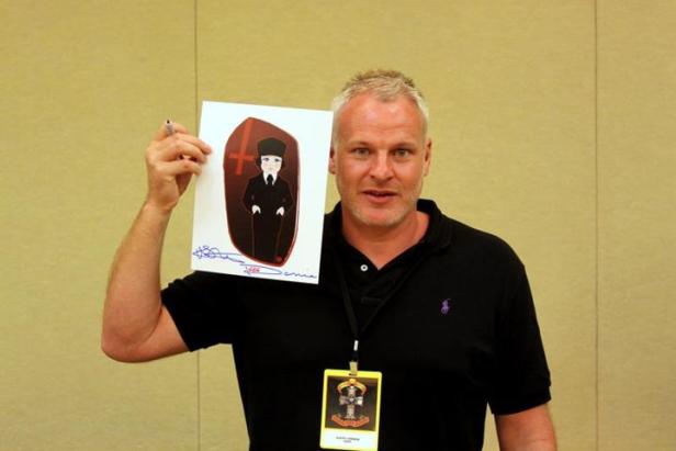 Ein Mann hält ein signiertes Bild einer Cartoonfigur mit Hut und umgedrehtem Kreuz hoch.