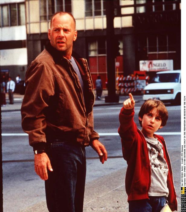 Bruce Willis steht mit einem Jungen, der auf etwas zeigt, auf einer Straße.