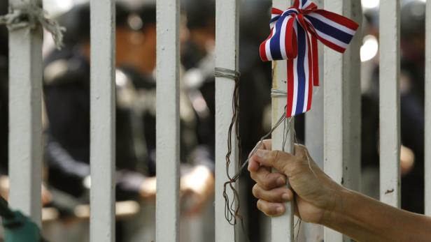 Eine Hand hält ein weißes Metallgitter, an dem eine Schleife in den Farben der thailändischen Flagge befestigt ist.