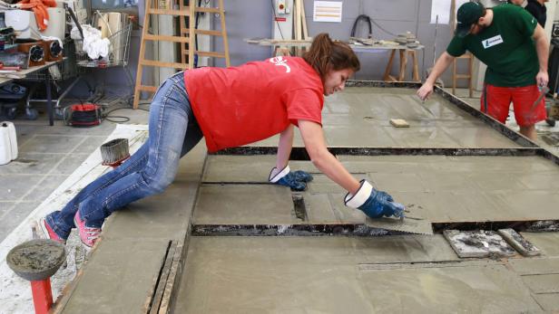 Eine Frau glättet Beton in einer Form, während ein Mann im Hintergrund arbeitet.