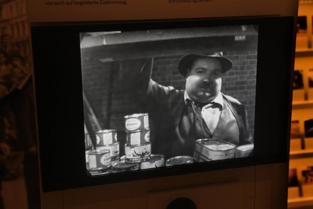 Ein Mann mit Hut steht vor einem Regal mit Konservendosen in einer Schwarzweißaufnahme des ORF.