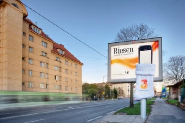 Drei Handysocke - Hello München/Gewista Rolling Board/Standort Wien Eine Werbetafel für „Riesen“ mit einem übergestülpten Socken auf einem Pfosten am Straßenrand.