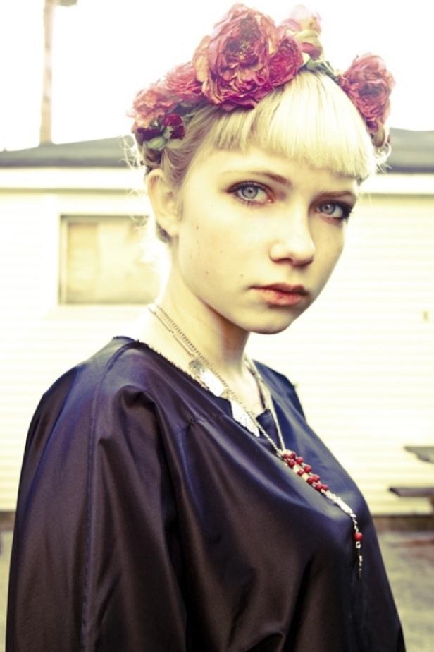 Eine junge Frau mit blonden Haaren und einem Blumenkranz auf dem Kopf blickt in die Kamera.