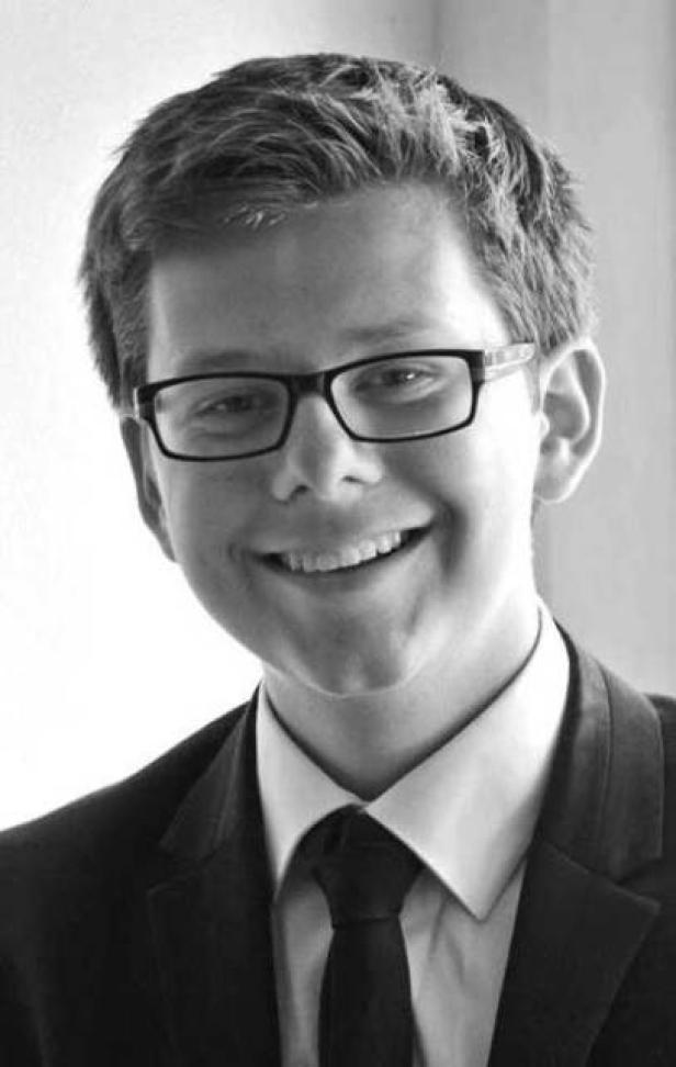 Ein lächelnder junger Mann mit Brille und Anzug in Schwarzweiß.