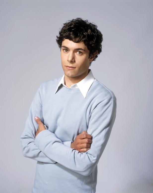 Adam Brody mit verschränkten Armen vor einem grauen Hintergrund.