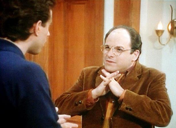 George Costanza spricht mit verschränkten Händen mit jemandem.