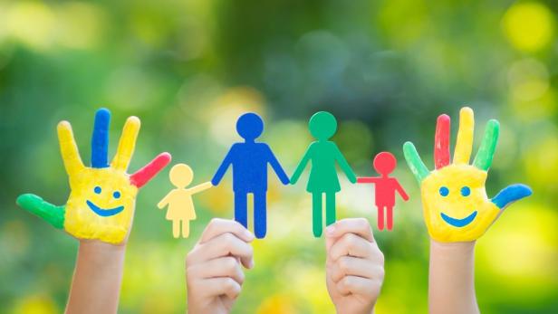 Kinderhände halten eine bunte Papierfamilie vor einem grünen Hintergrund.