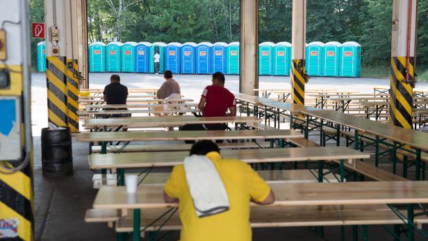 Mehrere Personen sitzen an Biertischen vor einer Reihe von Toilettenhäuschen.
