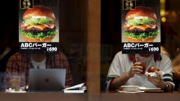 Zwei Personen sitzen in einem Restaurant, im Fenster hängen Burger-Werbeplakate.