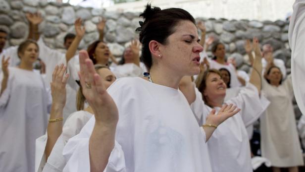 Eine Gruppe von Menschen in weißen Gewändern steht mit erhobenen Händen vor einer Steinwand.