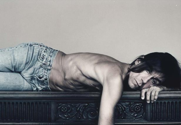 Iggy Pop liegt mit nacktem Oberkörper und Jeans auf einem verzierten Möbelstück.
