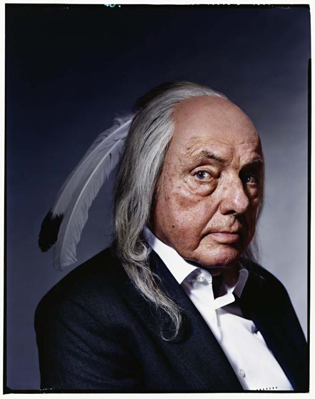 Ein älterer Mann mit einem Anzug und einer Feder im Haar blickt in die Kamera.