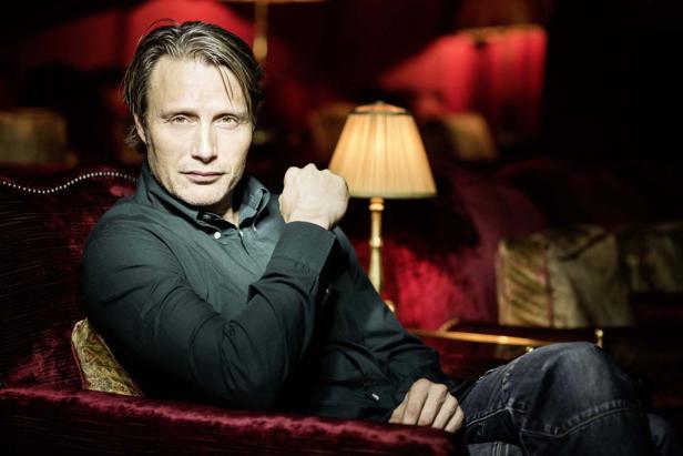 Mads Mikkelsen sitzt auf einem roten Sofa in einem eleganten Raum.