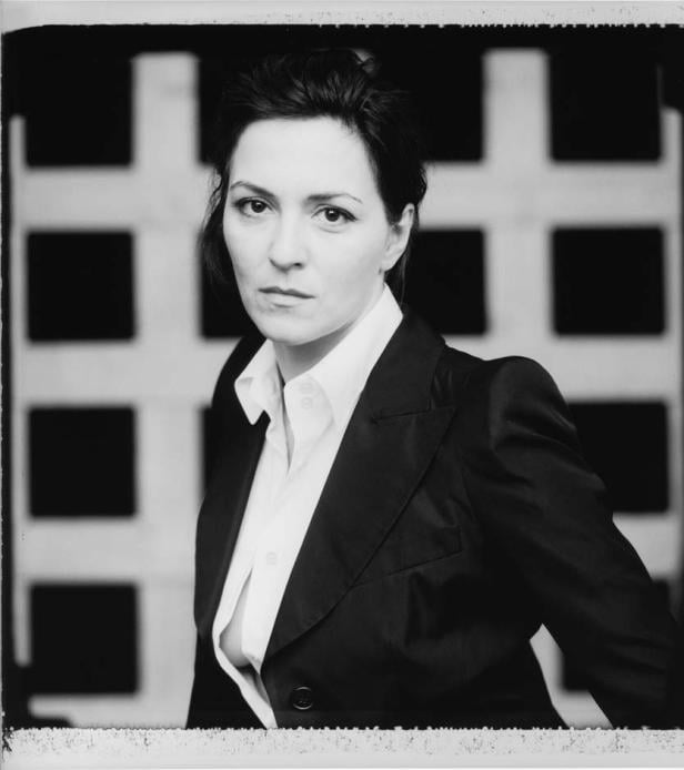 Schwarzweißporträt einer Frau in Blazer und weißem Hemd vor einem Gitter.