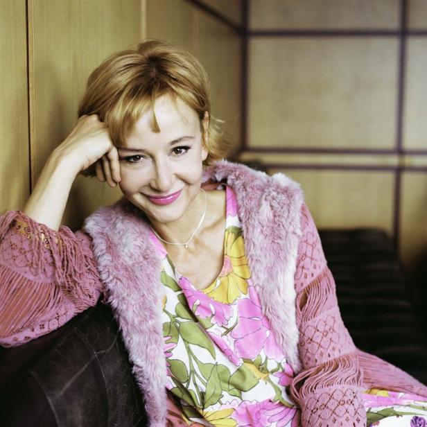 Eine lächelnde Frau mit kurzem blonden Haar, gekleidet in ein buntes Blumenkleid und eine rosa Jacke.