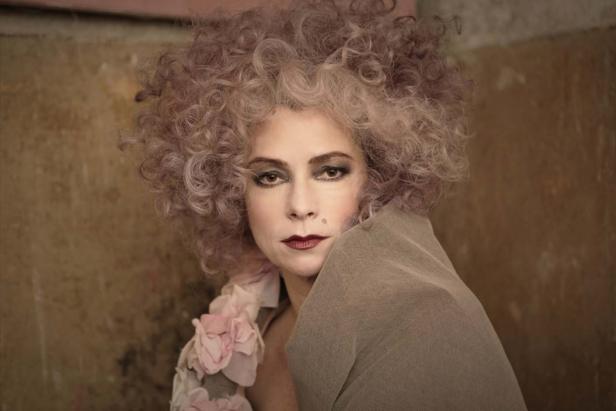 Eine Frau mit lockigem Haar und dunklem Lippenstift blickt in die Kamera.