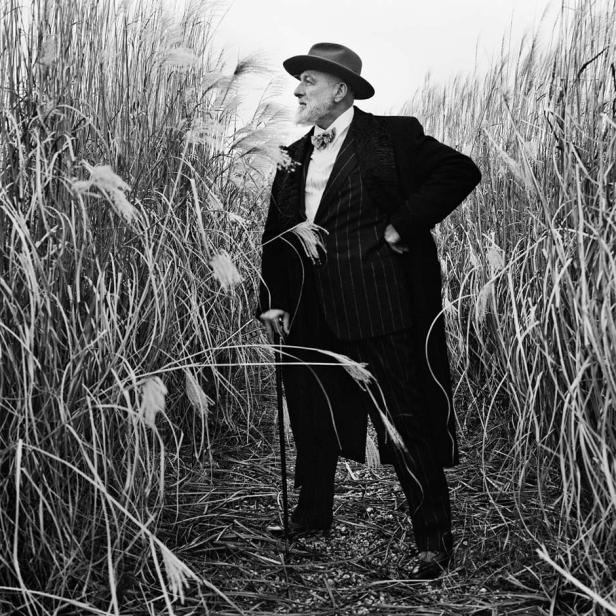 Ein älterer Mann mit Hut und Anzug steht mit einem Stock in hohem Gras.