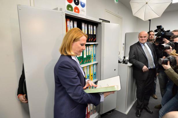 Eine Frau steht vor einem Aktenschrank und hält einen Ordner in der Hand, während sie von Reportern fotografiert wird.