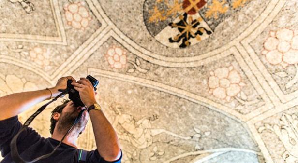 Ein Mann fotografiert eine kunstvoll bemalte Decke mit Wappen und floralen Mustern.