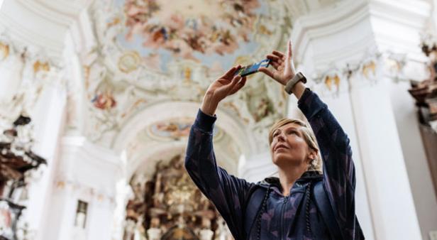Eine Frau fotografiert mit ihrem Smartphone ein Deckenfresko in einem Kirchenraum.
