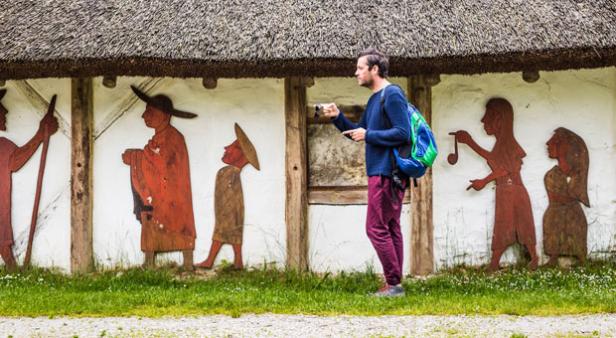 Ein Mann fotografiert hölzerne Figuren an der Wand eines Hauses mit Strohdach.