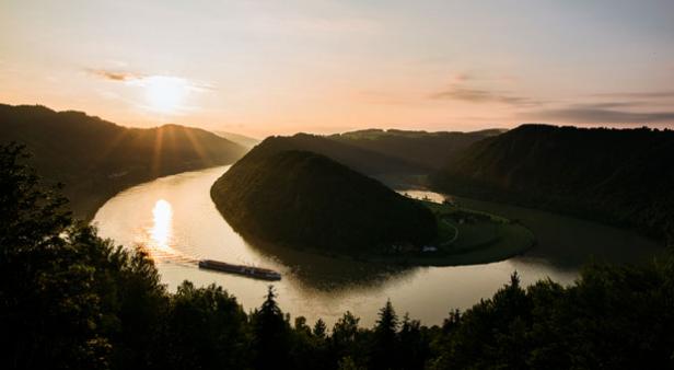 Ein Fluss schlängelt sich um einen bewaldeten Hügel bei Sonnenuntergang, ein Schiff fährt auf dem Wasser.