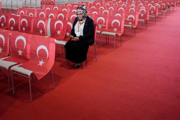 Eine Frau sitzt in einem Saal voller Stühle, die mit türkischen Flaggen geschmückt sind, und blickt auf ihr Handy.