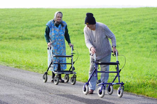 Zwei ältere Menschen gehen mit ihren Rollatoren auf einem Weg entlang.