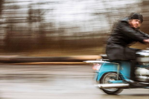 Ein Mann fährt mit einem blauen Motorroller auf einer Landstraße entlang.
