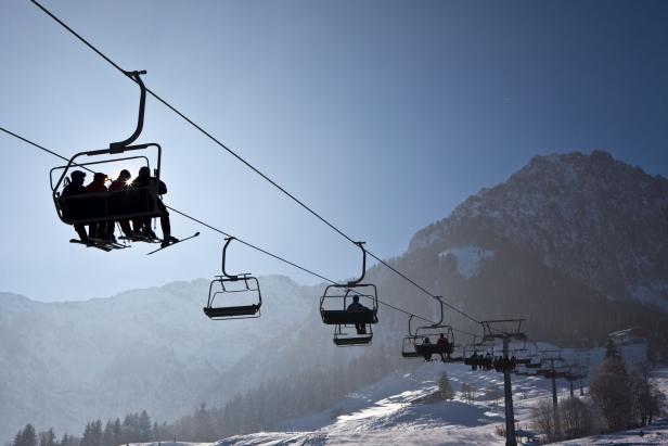 Mehrere Skifahrer sitzen in einem Sessellift vor einer verschneiten Berglandschaft.