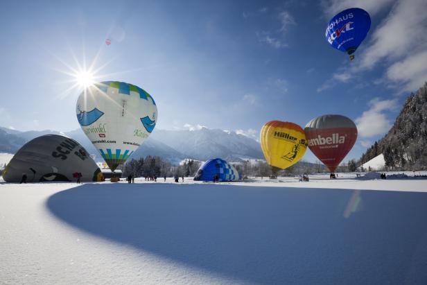 Mehrere Heißluftballons liegen auf einer schneebedeckten Fläche vor einer Bergkulisse.