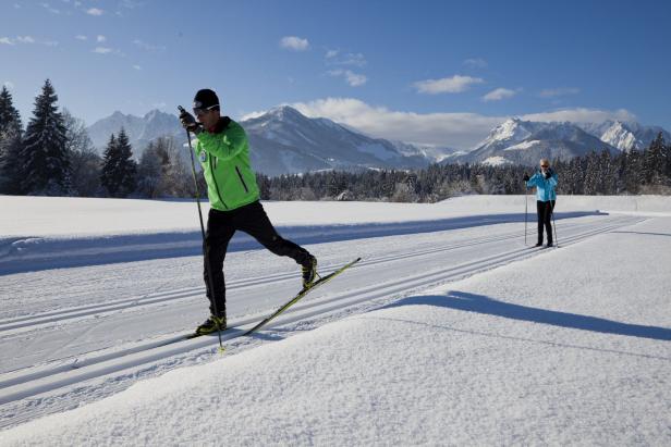 Zwei Personen laufen auf einer Loipe vor einem Bergpanorama Ski..