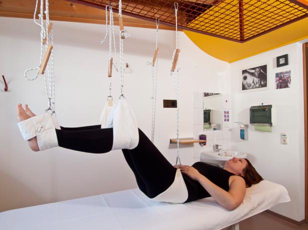 Eine Frau liegt in einem Therapieraum und wird mit Seilzügen behandelt.