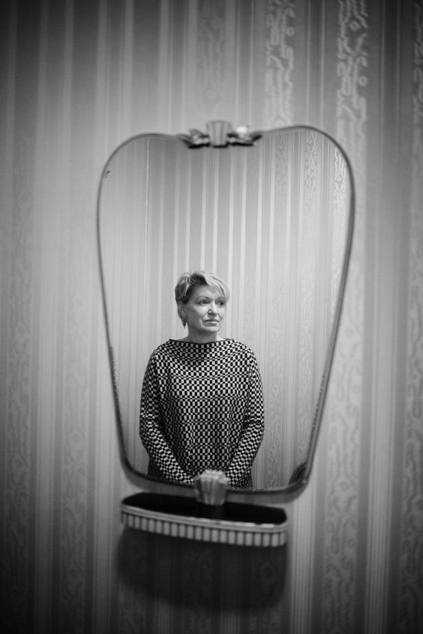 Eine Frau mit kurzem Haar steht vor einem Spiegel in einem Raum mit gestreifter Tapete.