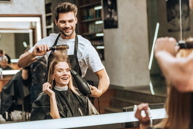 Ein Friseur föhnt und bürstet das Haar einer lächelnden Kundin.