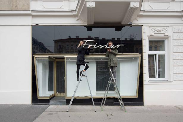 Zwei Personen montieren ein Leuchtschild mit der Aufschrift „Frisier“ an einem Gebäude.