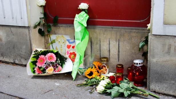 Blumen, Kerzen und eine Zeichnung liegen vor einer roten Tür als Gedenkstätte.