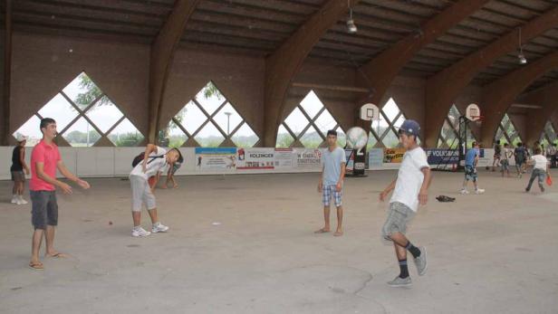 Mehrere Jugendliche spielen in einer Halle mit Basketballkörben und dreieckigen Fenstern.