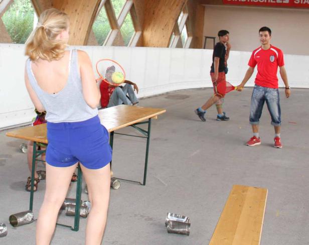 Junge Leute spielen mit Dosen und Tennisschlägern in einer Halle.