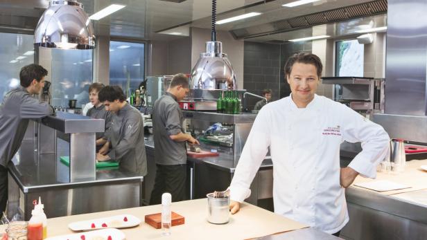 Ein Koch steht in einer modernen Restaurantküche, während andere Köche im Hintergrund arbeiten.