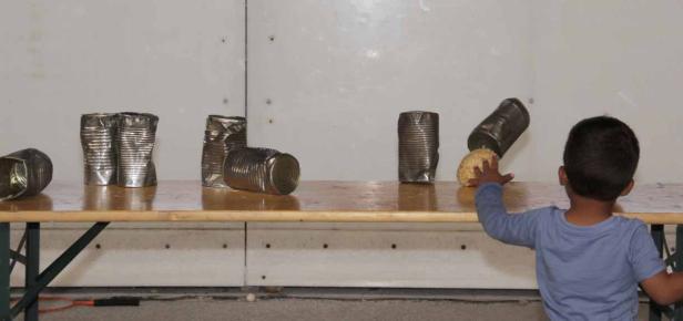 Ein Kind spielt mit einer Kartoffel und umgestoßenen Konservendosen auf einem Tisch.