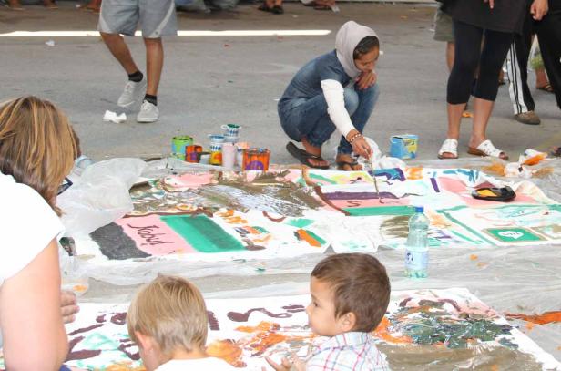 Eine junge Frau mit Kopftuch malt mit anderen Kindern an einem großen Gemeinschaftsbild.