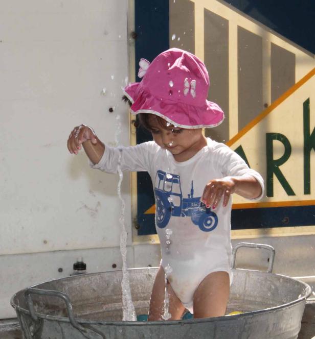 Ein kleines Kind mit rosa Hut spielt mit Wasser in einer Zinkwanne.