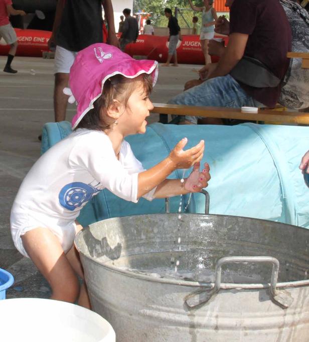 Ein kleines Mädchen mit rosa Hut wäscht sich die Hände in einem Zinkeimer.