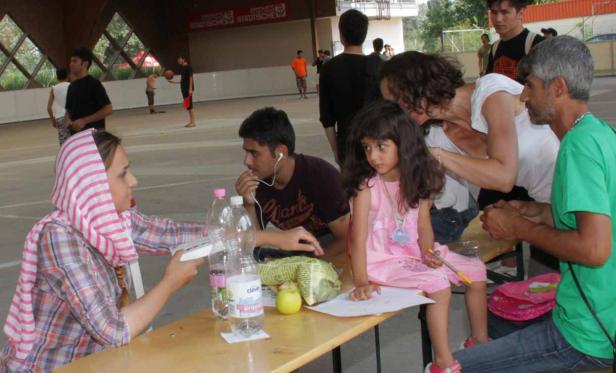 Eine Gruppe von Menschen sitzt an einem Tisch, während im Hintergrund andere auf einem Sportplatz aktiv sind.