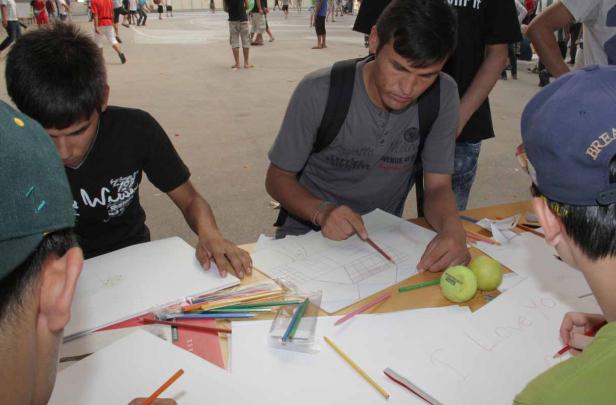 Mehrere junge Männer sitzen an einem Tisch und zeichnen mit Buntstiften auf Papier.