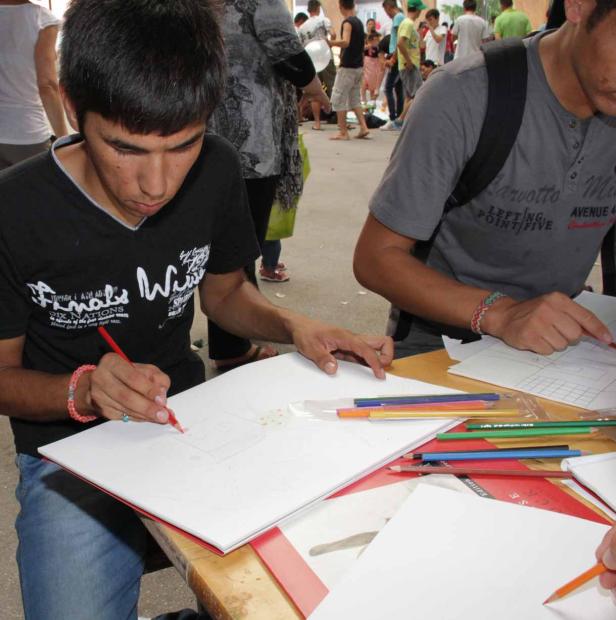 Zwei junge Männer zeichnen mit Buntstiften auf Papierbögen im Freien.
