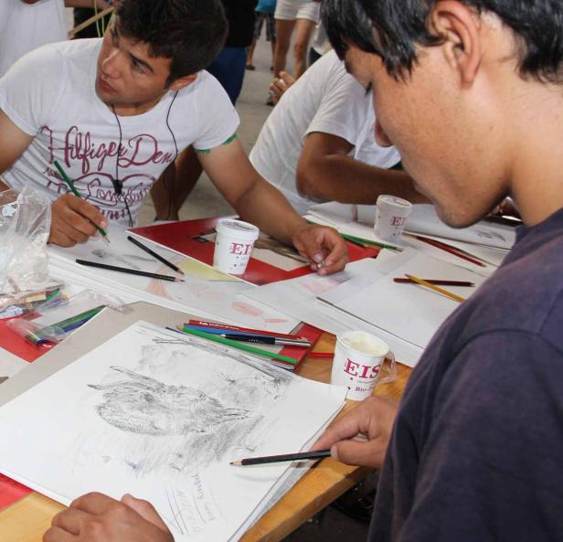 Zwei junge Männer zeichnen an einem Tisch im Freien mit Bleistiften und Buntstiften.